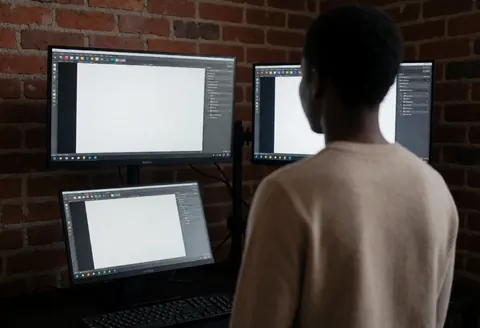 Person reviewing color monitors in a room with a brick wall, illuminated by the light from the screens.
