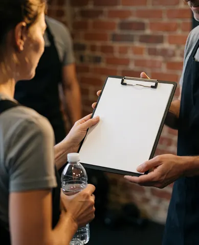 Fitness trainer presenting a workout plan on a clipboard to a client in a gym with a brick wall background