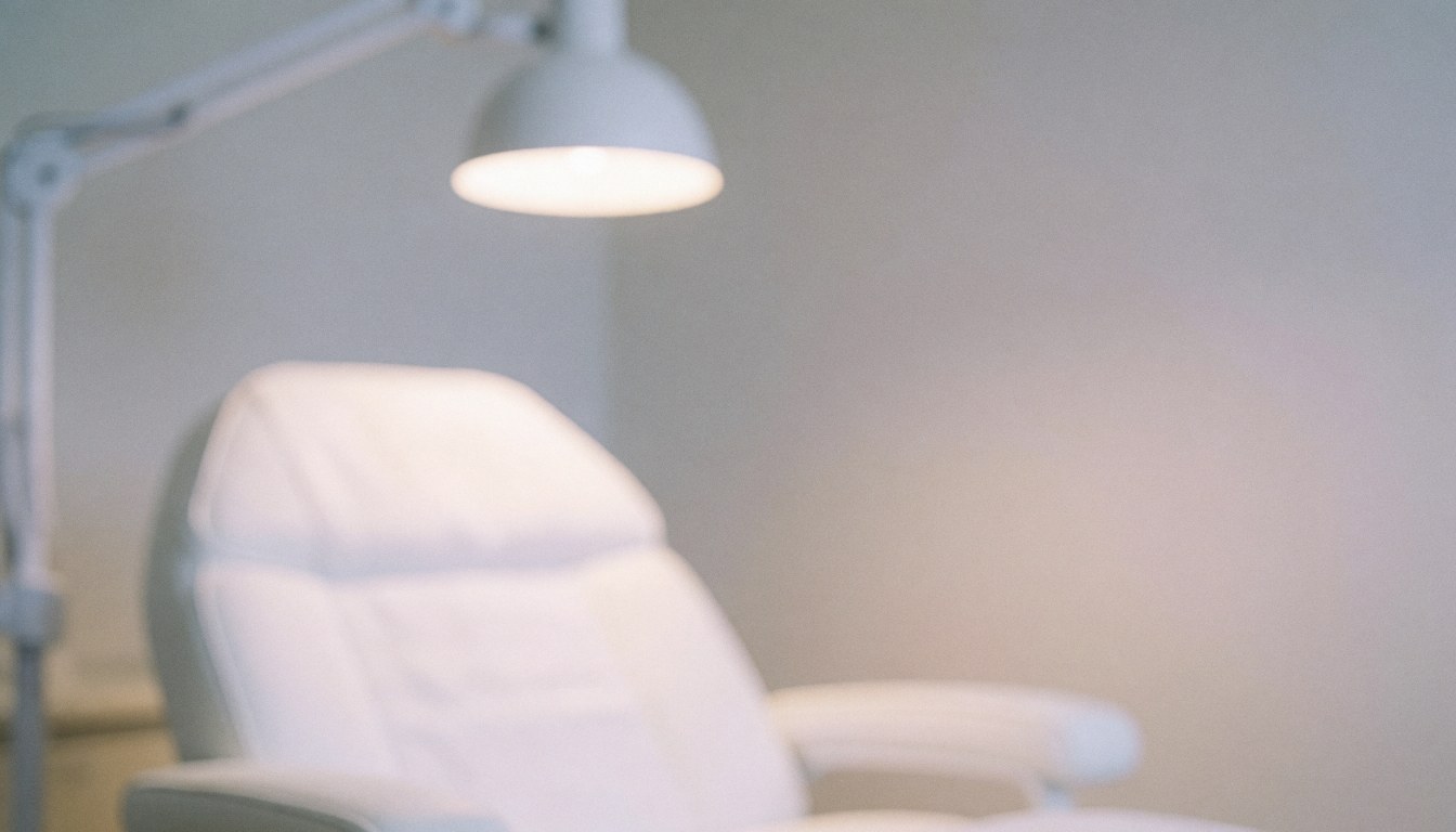 Blurred background of a treatment room featuring a white chair and wall lamp with serene minimalist aesthetic, pastel colors and warm lighting.