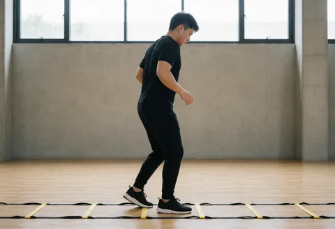 A man in black athletic wear performs agility ladder drills on a wooden floor in a gym with concrete walls and natural light.