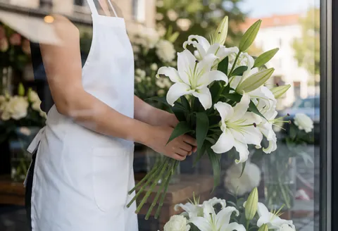 A florist adjusts a bouquet of white lilies with green leaves, viewed from outside through the storefront window.