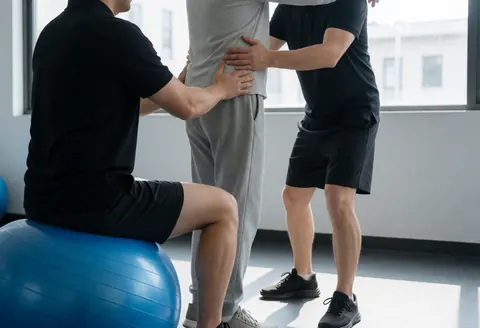 A personal trainer supports a client performing an exercise on a blue stability ball in a gym with gray walls.