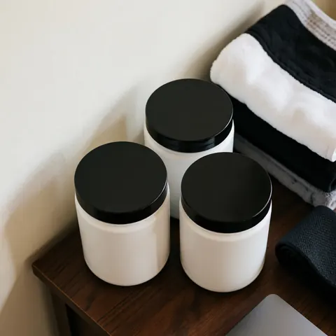 Three white supplement containers with black lids arranged on a dark wooden surface alongside folded sports towels in a fitness setting
