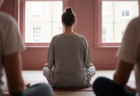 Yoga instructor sitting in lotus pose in a room with pink walls and natural light, viewed from behind during a class