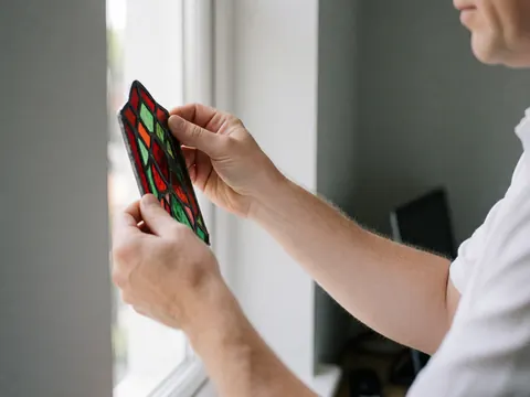 Close-up of glazier hands positioning red and green patterned glass pieces against natural light in a workshop setting