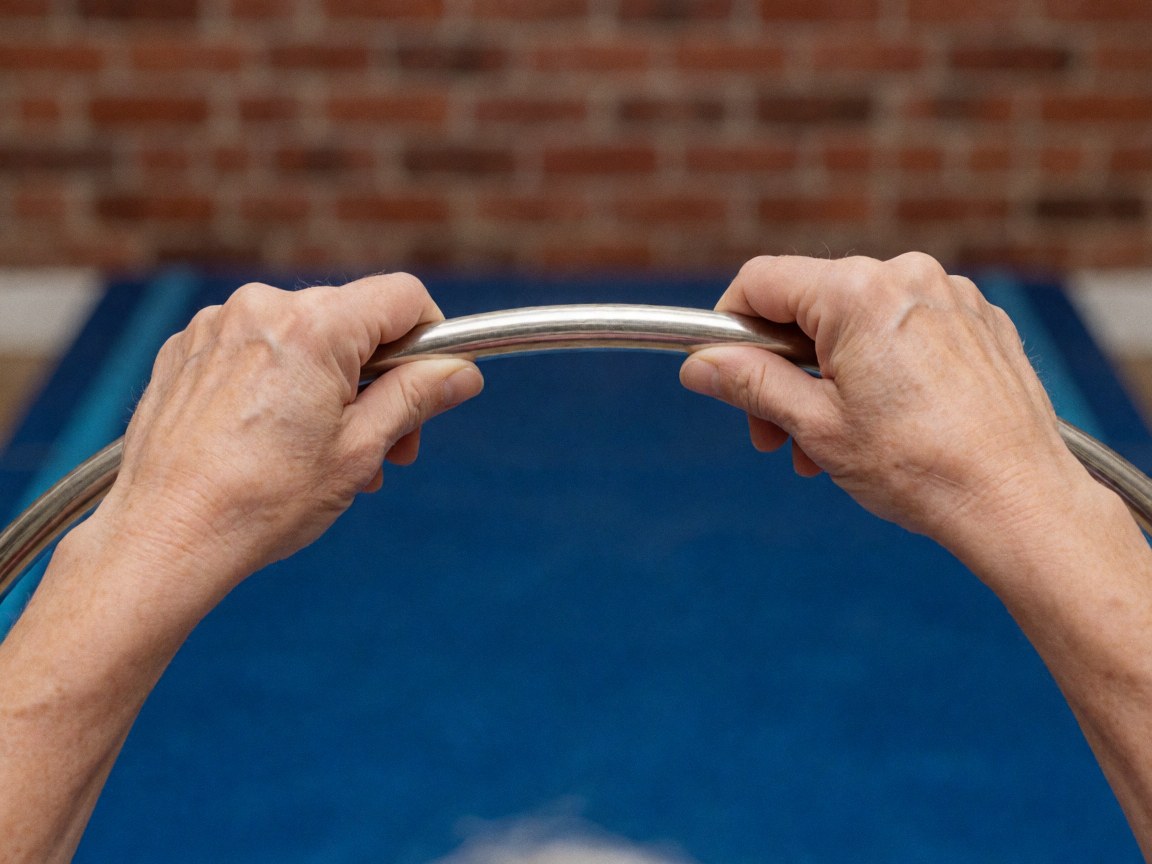 Adult hands gripping a metal gymnastics hoop against a blue mesh background with a red brick wall behind it