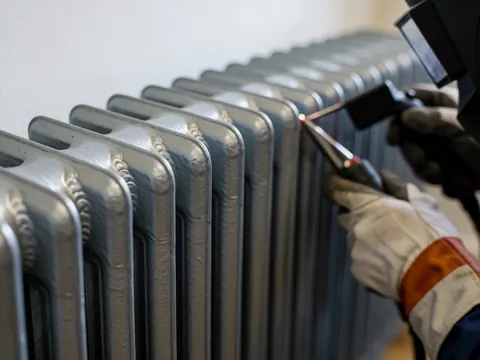 Close-up of a gloved hand using a welding tool to repair an aluminum radiator, with visible weld marks and work tools in a boiler repair shop.