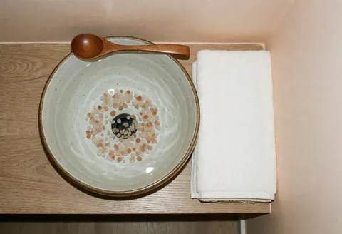 Ceramic bowl filled with pink salt and a wooden spoon placed next to a folded white towel on a clean surface for foot therapy.