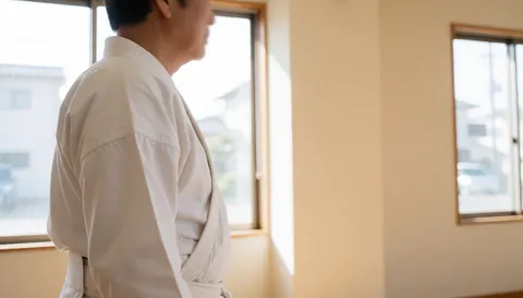 Side profile of an adult in a white martial arts uniform standing in a dojo with beige walls and natural light