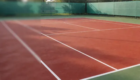 Empty red clay tennis court with white boundary lines and a green fence, suitable as a background for tennis instructor content