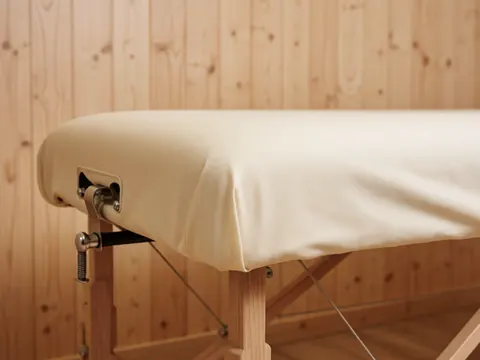 Close-up of a fixed metal latch on the corner of a massage table covered with cotton fabric, set against a natural wood wall with soft lighting.