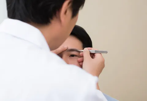 Close-up of a specialist using tweezers to perform microblading on a client's eyebrow against a beige background with bright lighting.