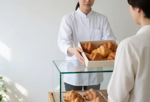 A baker in a white uniform serves a box of croissants from a glass display case in a bright bakery with white walls.