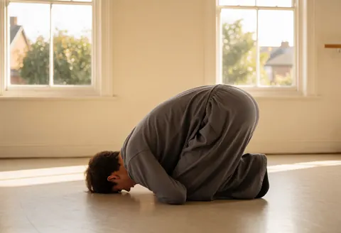 A dance instructor executes a floor movement on a wooden floor in a bright studio with light walls and a ballet barre.