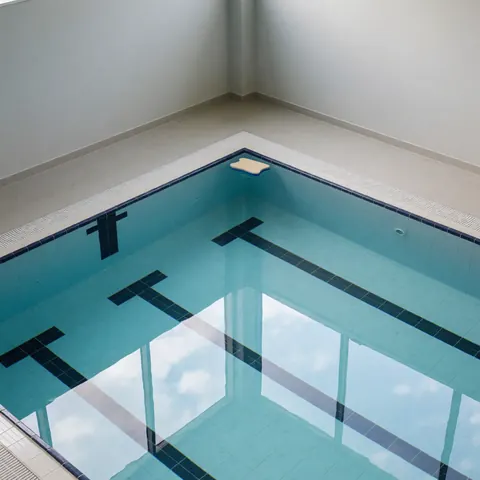 Indoor swimming pool featuring lane lines and a floating kickboard against light gray walls with clear water reflecting the sky.