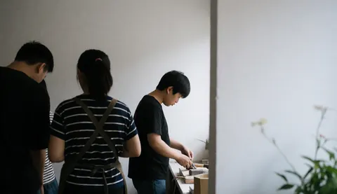 Three individuals observe a carpenter working with tools in a workshop with white walls