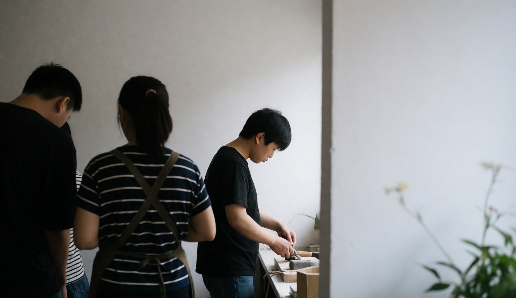 Three individuals observe a carpenter working with tools in a workshop with white walls