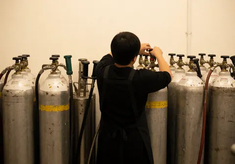 Person in black apron adjusting a gas tank valve among other tanks in a warm white-walled workshop for event decorators.