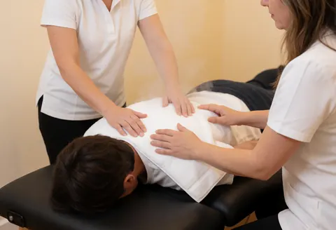 Two therapists applying steam therapy to a client's back on a massage table in a warm professional setting