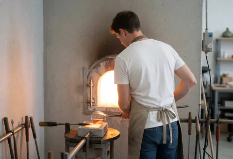 Glassblower shaping molten glass using tools and silicone molds in a workshop with concrete walls.