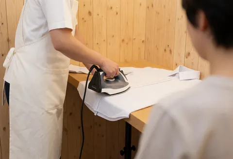 A tailor wearing a white apron irons a shirt seam on a wooden board against a natural paneled wall background