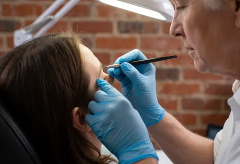 A microblading artist uses a manual tool to apply precise strokes to a client's eyebrows while wearing blue gloves in a studio with a brick wall.