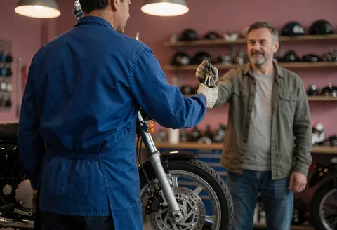 Mechanic handing keys to a customer in a motorcycle repair shop with a front wheel and helmet shelves visible