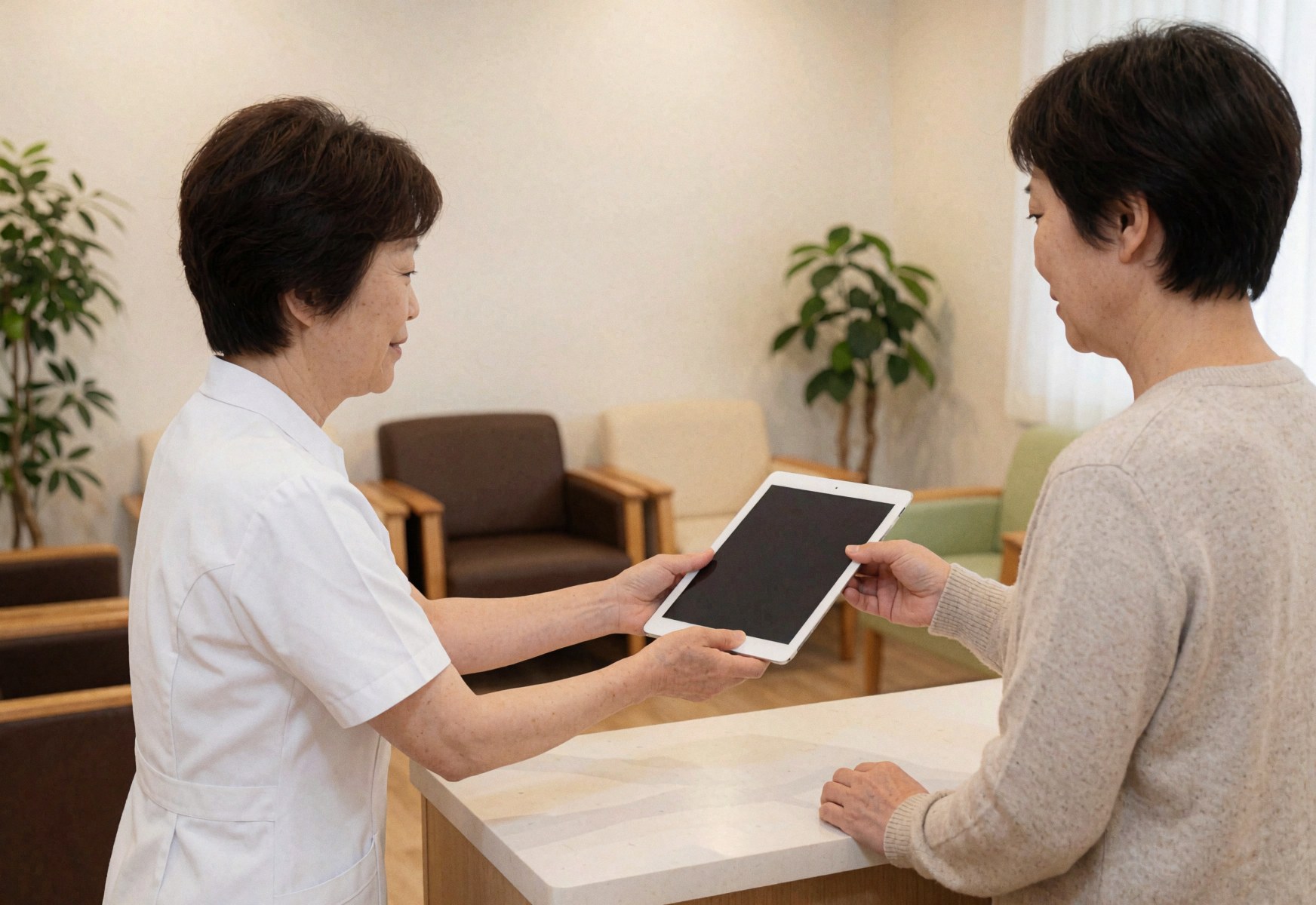 Therapist in white uniform presents digital tablet to client in a clean, modern reception area with chairs and plants.