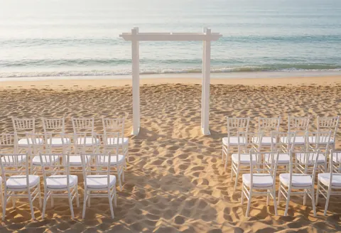 A white arch on the beach with white chairs, ceremony scene by the sea, soft morning light
