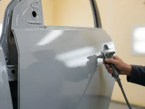 Close-up of a hand using a spray gun to apply gray base coat paint to a car door in an auto body shop.