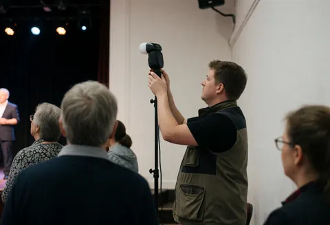 Professional photographer adjusting a flash unit during an event with an audience and stage lighting in a professional setting.