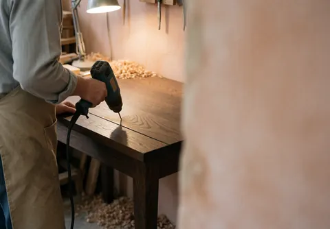 Carpenter repairing a wooden table with an electric drill, wood shavings, and desk light in a workshop with terracotta stucco walls.