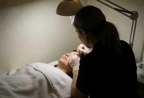Aesthetician wearing gloves applies facial serum to a client resting on a white towel in a warm-toned room