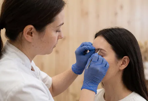 Close-up of hands in blue gloves using tweezers to apply false eyelashes to a client's closed eye in a salon with light wood walls