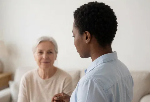 A hypnotherapist speaking with a seated patient in a bright white room with soft lighting and plain walls during a professional session