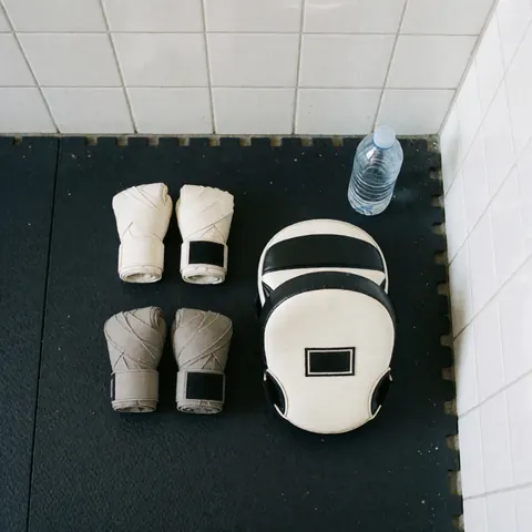 Boxing gloves, hand wraps, and training masks arranged on a black rubber mat against a white tiled wall, suitable for boxing instructor business use.