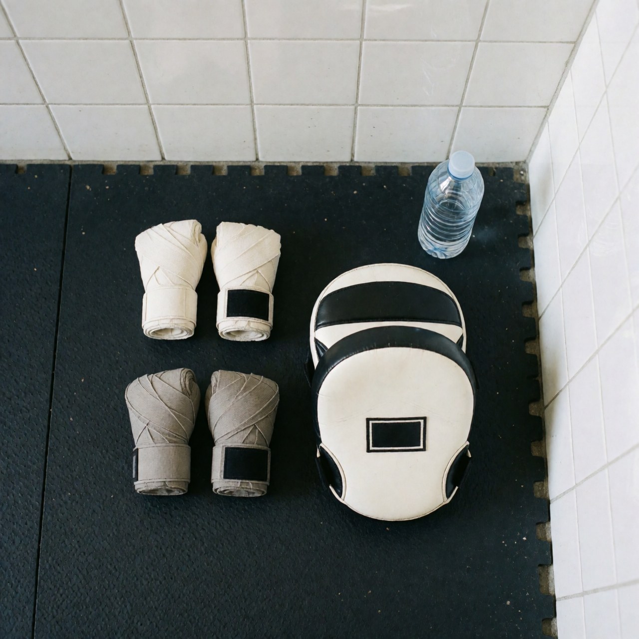 Boxing gloves, hand wraps, and training masks arranged on a black rubber mat against a white tiled wall, suitable for boxing instructor business use.