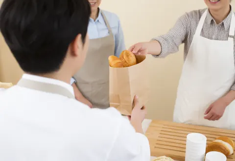A bakery employee hands a package of fresh bread to a smiling customer in a warm shop setting.
