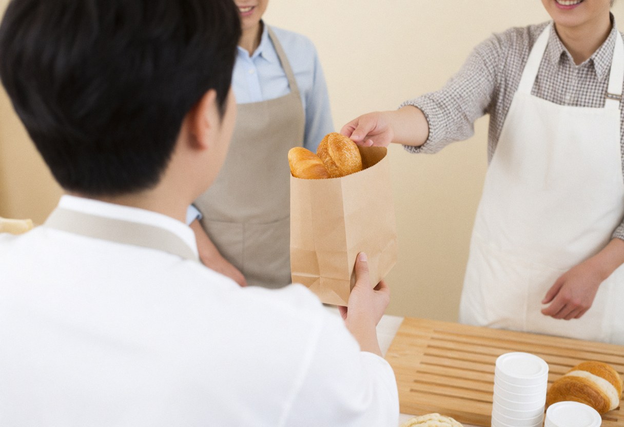 A bakery employee hands a package of fresh bread to a smiling customer in a warm shop setting.