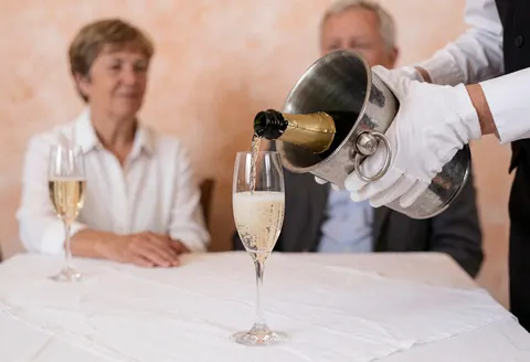 A server pours champagne into a flute on a white tablecloth during a banquet event with guests seated in the background