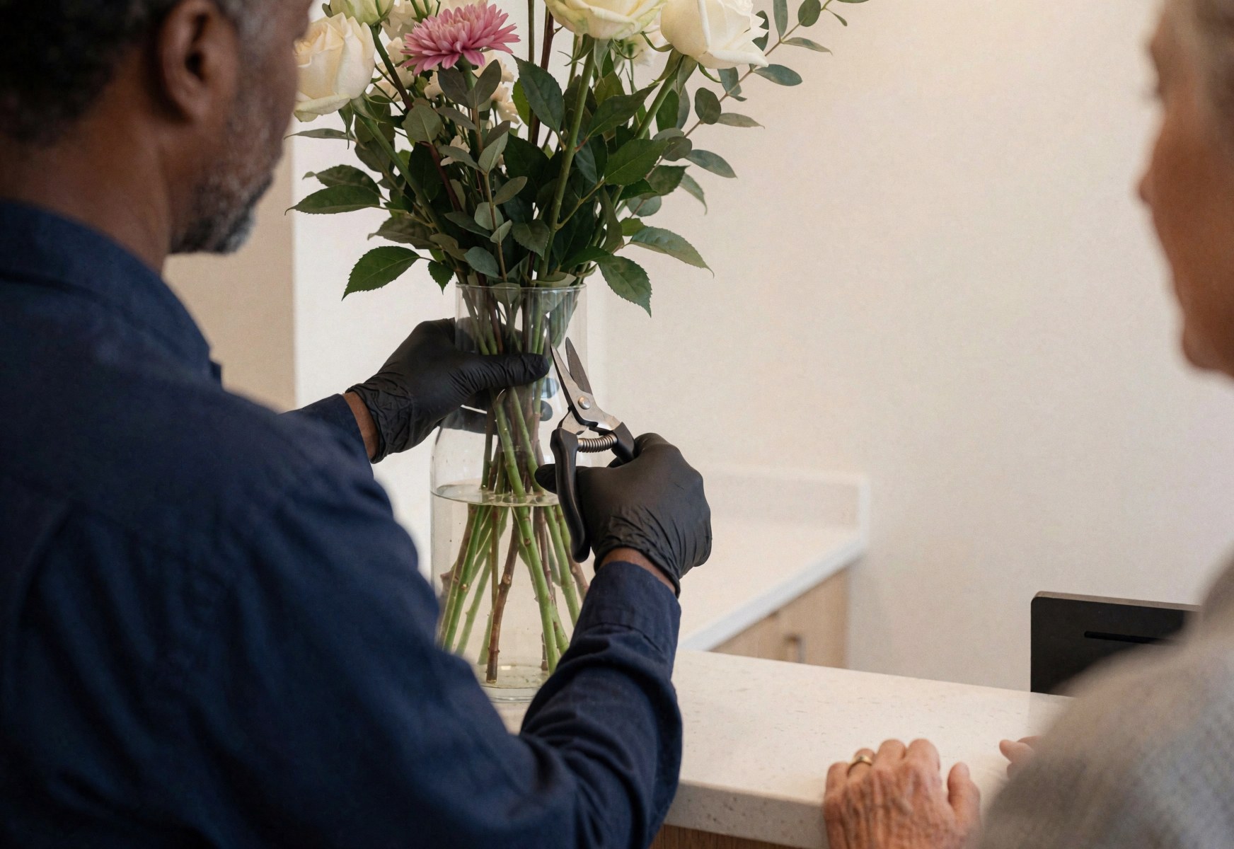 A florist wearing gloves cuts rose stems at a glass vase on a reception counter with a client nearby