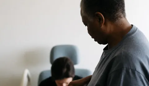 A professional geriatric massage therapist providing a seated massage service in a bright room with natural light and a white wall background.