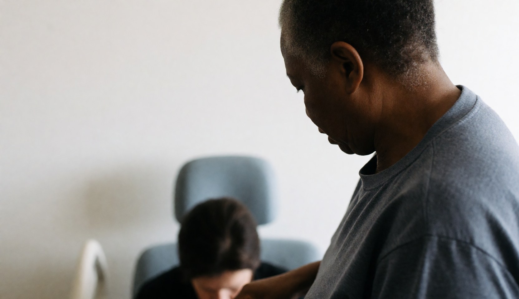 A professional geriatric massage therapist providing a seated massage service in a bright room with natural light and a white wall background.