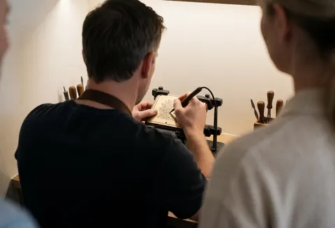 Close-up of hands carving a design into a wooden plaque with tools in a workshop with white walls