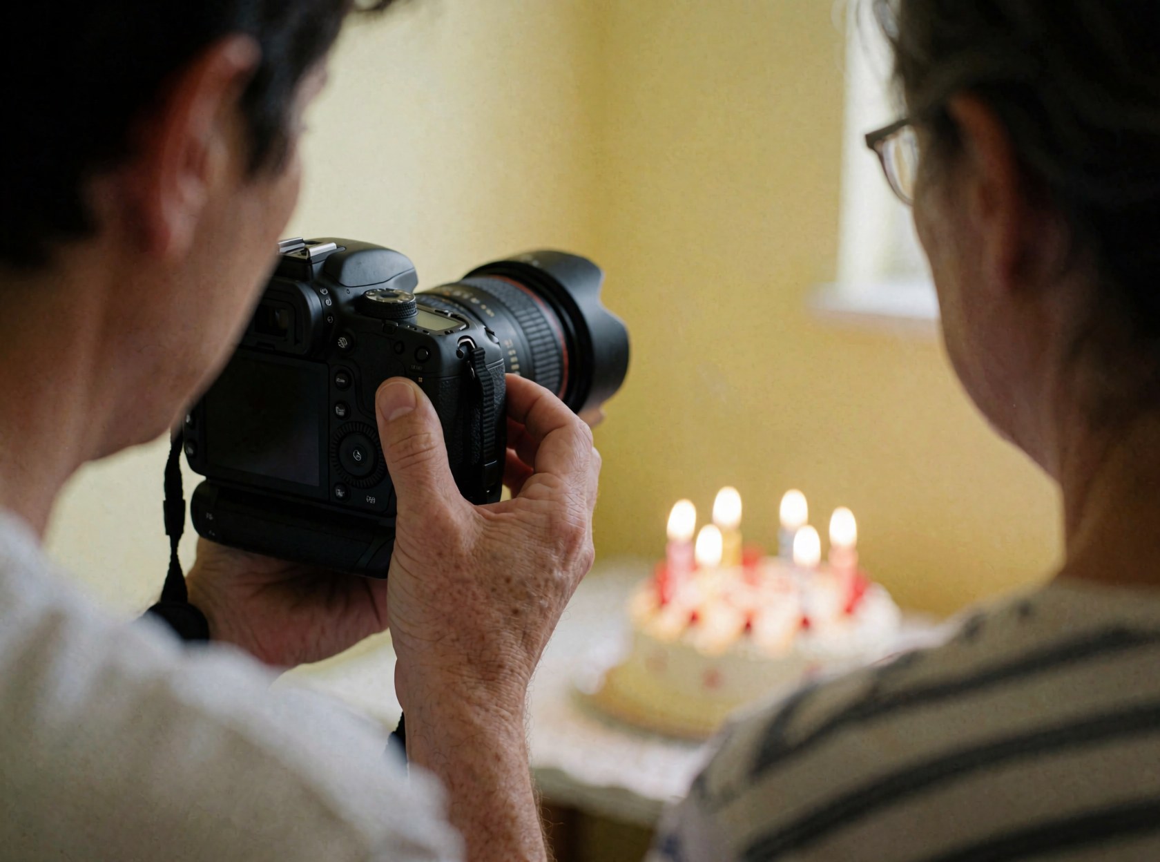 A photographer's hand adjusts a camera lens while standing in front of a birthday cake with lit candles in a room with yellow walls.