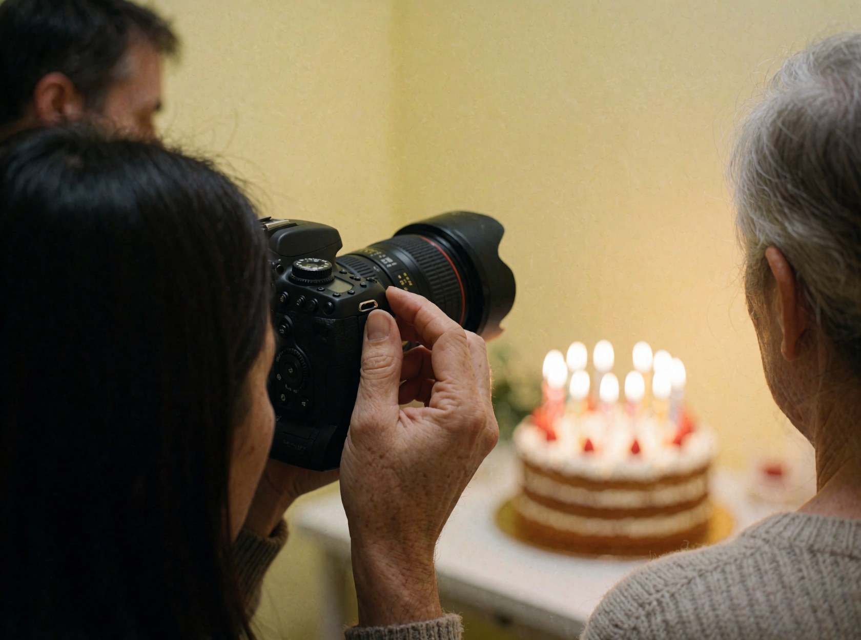 A hand adjusts the focus ring on a dslr camera pointed at a birthday cake with lit candles against a yellow wall.