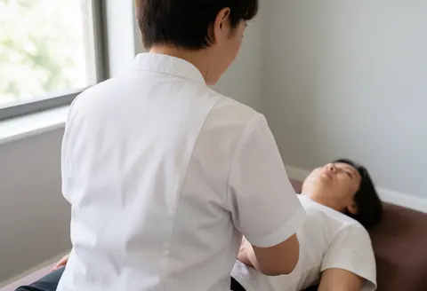 Shiatsu practitioner applying pressure to a client's abdomen while she lies on a mat in a naturally lit room with warm gray walls