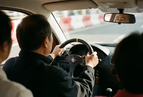 A driver's hand on the steering wheel and side mirror visible in a racing car on an asphalt track with red and white barriers