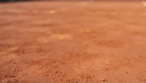 Out of focus red clay tennis court surface with natural lighting, serving as a textured backdrop for sports service text
