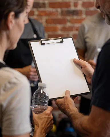 A fitness trainer holds a white clipboard while standing next to a client in a gym with a brick wall and water bottles.